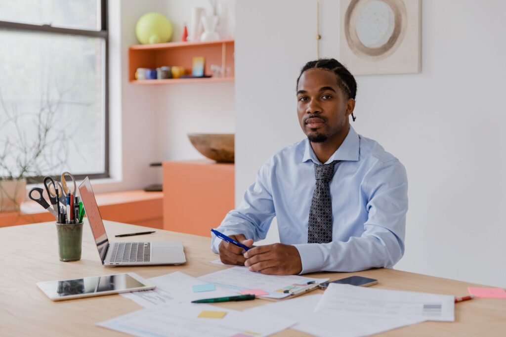 A professional businessman with a beard and braided hair sits at his desk with a laptop and documents, working diligently.
