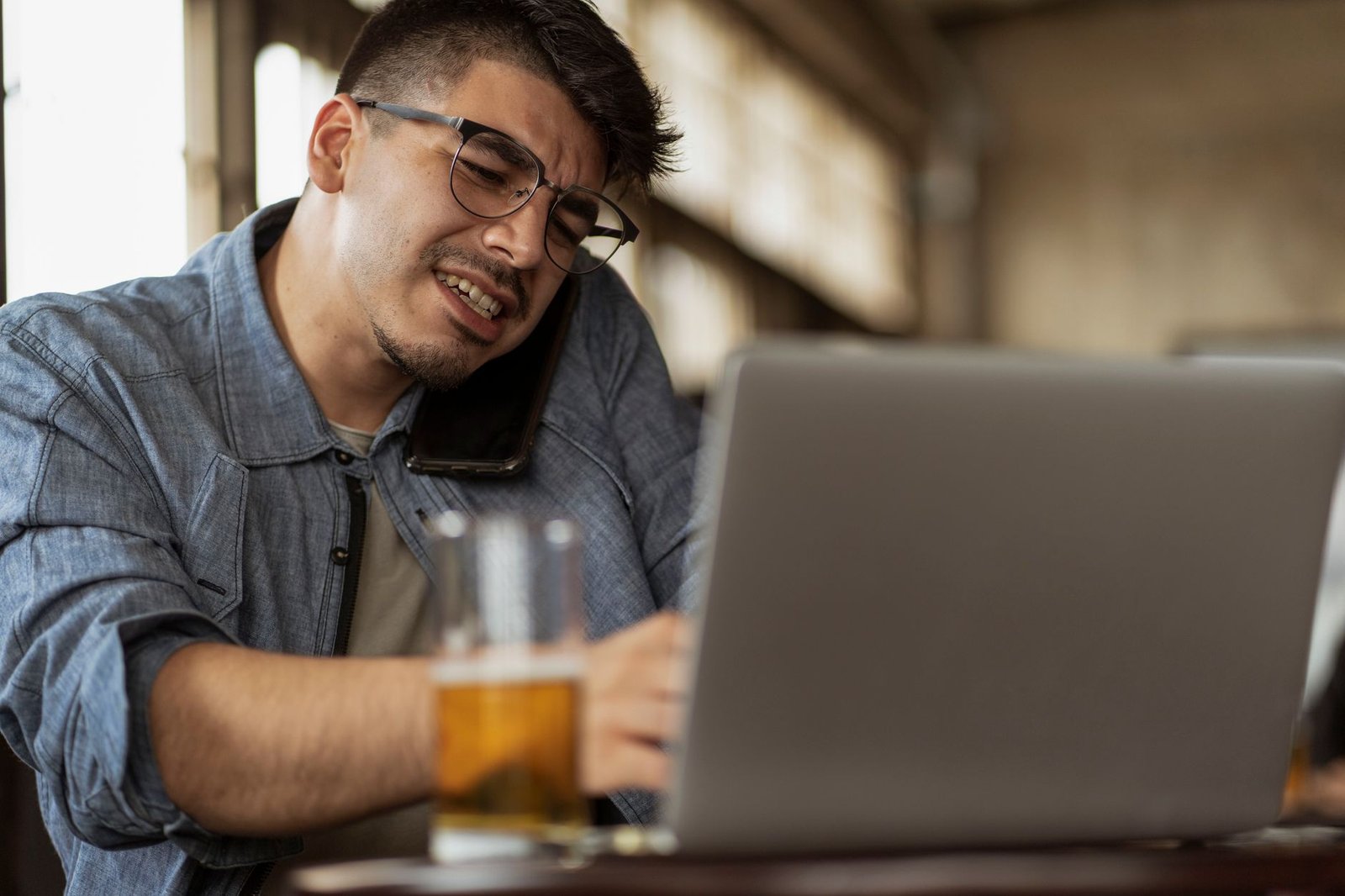 Young man multitasks, talking on phone and working on laptop in a cozy cafe setting.